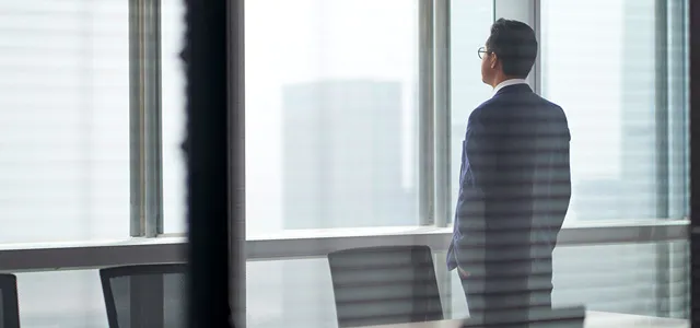 Business professional in a suit standing by office window, looking at city skyline in a modern workspace.